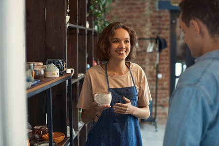 Smiling attractive clerk in apron standing at shelf and selling ceramic cup to man in pottery storeの写真素材