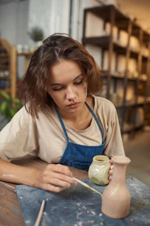 Concentrated young Caucasian potter sitting at table and painting vase while working on its design...