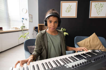 Serious African-American schoolgirl sitting by piano keyboard during repetition and looking at youの写真素材
