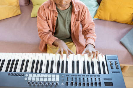 Contemporary multiracial schoolboy pressing keys of piano keyboard while sitting on couch with many cushions at homeの写真素材