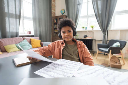 Happy cute African schoolboy with papers sitting by table during lesson in home environment