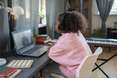 Cute schoolgirl in headphones looking at teacher on laptop screen while sitting by table in home...