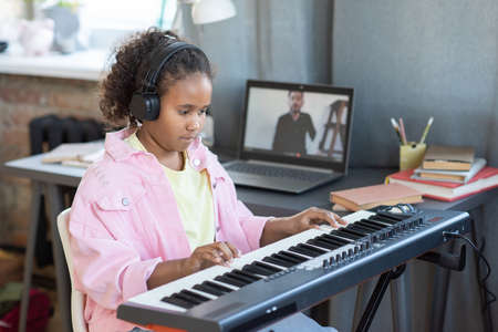 Pretty schoolgirl in headphones playing piano keyboard while listening to advice of her teacher during online lessonの写真素材