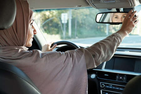 Happy young Muslim businesswoman in hijab looking in mirror while sitting by steering wheel and driving carの写真素材