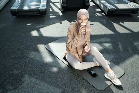 Young restful sportswoman drinking water after exercising while sitting on mat in large contemporary fitness centerの写真素材