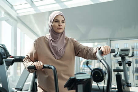 Young sportswoman in hijab holding by handles of sports equipment while exercising in modern fitness centerの写真素材