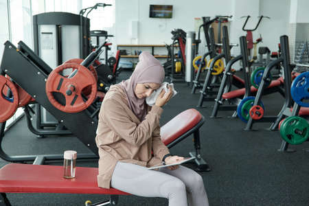 Young restful sportswoman touching her wet face by towel while sitting on exercise equipment after...