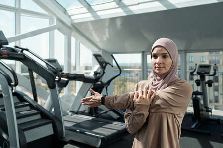 Young sportwoman in hijab doing physical exercise for arms while standing in gym among sports equipmentの写真素材