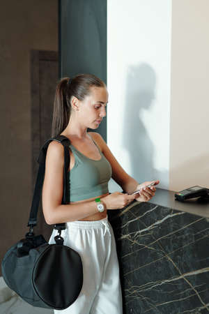 Brunette girl in activewear looking at smartphone screen while standing by reception counter in...