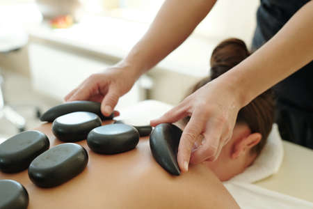 Part of body of young female with hot spa stones on her back and professional masseuse holding two of themの写真素材