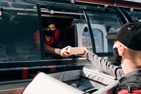 Young sales clerk in protective mask passing packed box with fast food to delivery service staffの写真素材