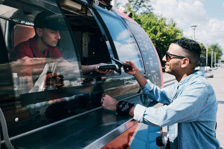 Young man buying fast food in street truck and holding smartphone over terminal during contactless...
