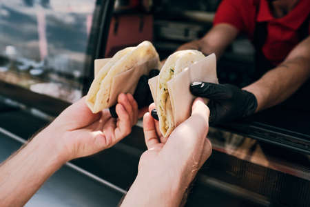 Hands of young man taking tasty snacks wrapped into paper held by gloved worker of food truckの写真素材