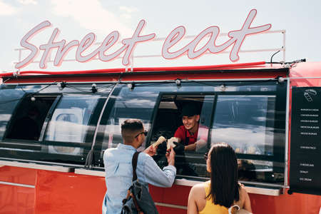 Happy young man in uniform passing two snacks to one of customers buying street foodの写真素材