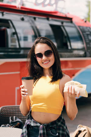 Happy young brunette female in sunglasses holding burger and drink bought in street food truckの写真素材