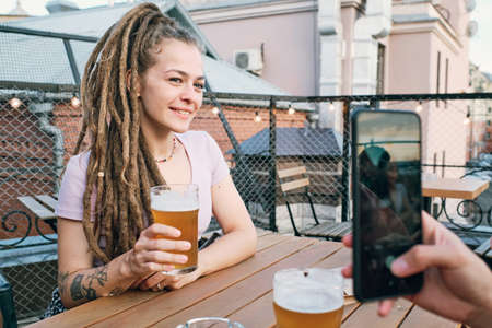 Happy girl with dreadlocks holding glass of beer while sitting by table and posing for friend with smartphoneの写真素材