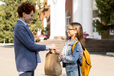 Young woman passing paperbag with snack to her little daughter against school building