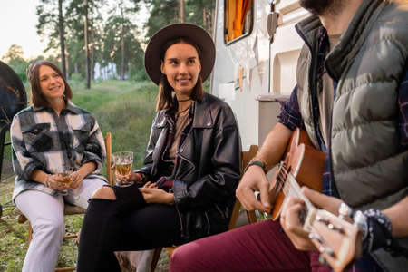 Two happy girl with drinks listening to young man playing guitar by house on wheels in the...