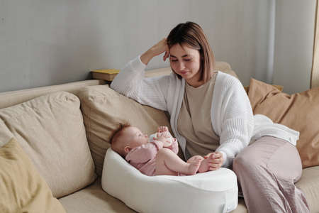 Happy young mother sitting on couch next to her adorable baby daughter with bottle of milk