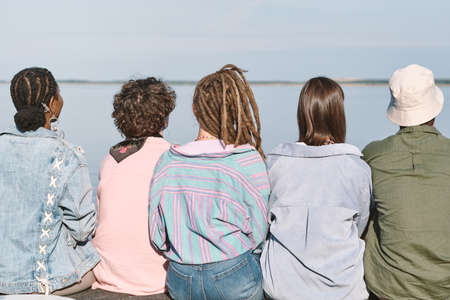 Rear view of young intercultural friends in casualwear sitting in row in front of watersideの写真素材