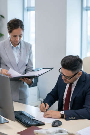 Mature employer signing documents by workplace while his secretary standing near byの写真素材