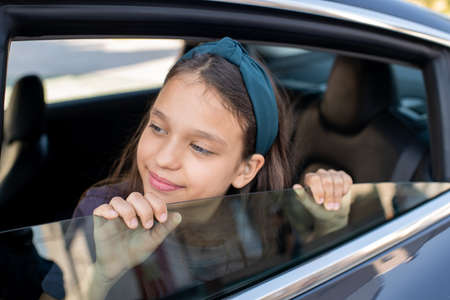 Adorable schoolgirl looking out of window of electric car while sitting insideの写真素材