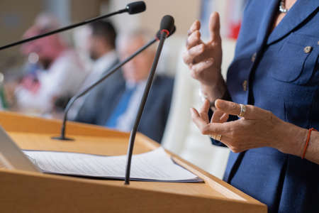 Close-up of unrecognizable female politician in rings standing at rostrum with microphones and clipboard while addressing conferenceの写真素材