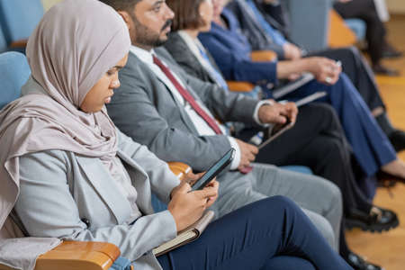 Concentrated Muslim nusinesswoman in hijab using smartphone while sitting at business conferenceの写真素材