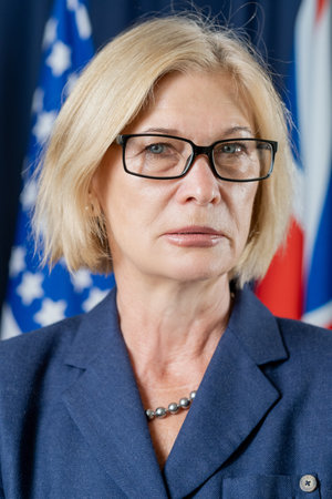 Portrait of serious mature blond-haired female politician in eyeglasses standing against flags