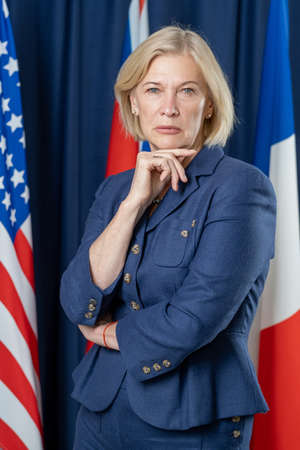 Portrait of serious mature female politician in formal suit holding hand on chin against national flagsの写真素材