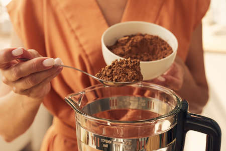 Hands of female putting spoon of cocoa powder into electric blender before mixing it with other ingredientsの写真素材