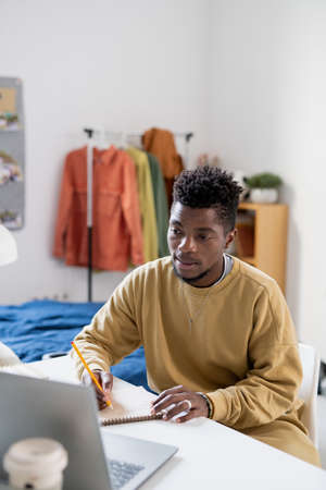 African guy in casualwear looking at laptop display while making notes during online lesson at homeの写真素材