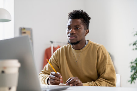 Tense young man with pencil looking at laptop screen while making notes and listening to teacher...