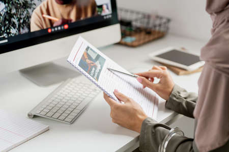 Hands of young contemporary teacher pointing at document while sitting in front of computer with her...