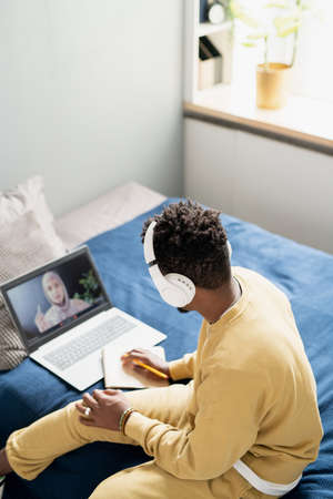 Young student in headphones and homewear listening to teacher and making notes while sitting on bed during online lessonの写真素材