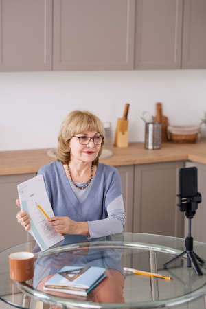 Happy blond mature teacher pointing at document while explaining information to online audience...