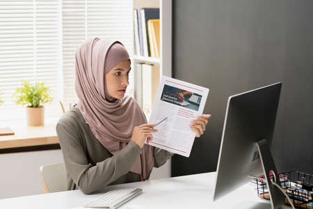 Confident teacher of economics pointing at financial document while sitting in front of computer...