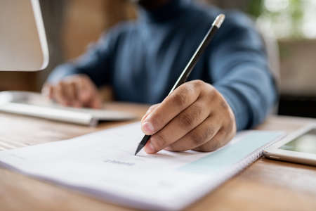 Hand of man holding pencil over document while sitting in front of computer and making notes during...