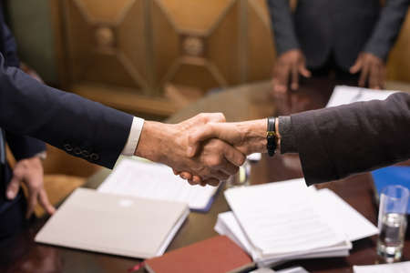 Hands of two mature successful business partners handshaking over table with signed documents after negotiationの写真素材