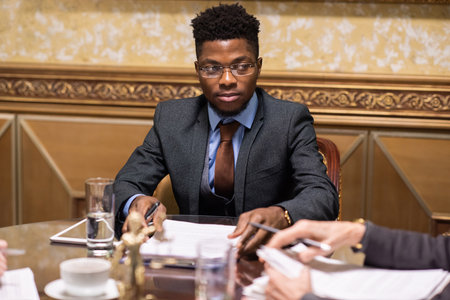 Young serious businessman in eyeglasses and elegant suit looking at colleague at meeting while...