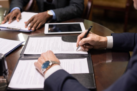 Hands of mature lawyer signing contract or other juridical document while sitting by table against...
