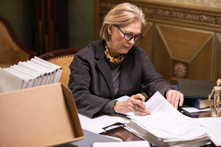 Blond mature woman in eyeglasses and formalwear looking for particular document while working with...