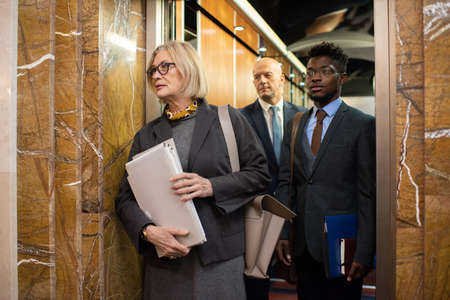 Group of contemporary intercultural co-workers with documents waiting for elevator while standing in...