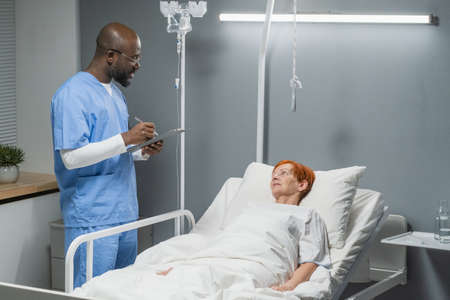 African male doctor writing prescription for his senior patient while she lying on the bed in the hospital wardの写真素材