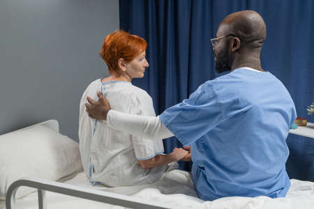 Rear view of African male doctor in uniform embracing and talking to mature woman while they sitting on the bed in the wardの写真素材