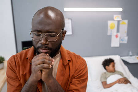 Close-up of African father with closed eyes praying for his little son while he lying in hospital bed behind himの写真素材