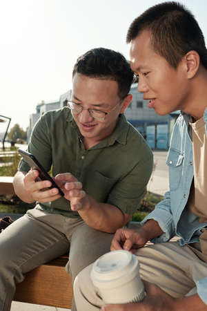 Small group of Chinese businessmen discussing photo or video in smartphone while resting in urban...