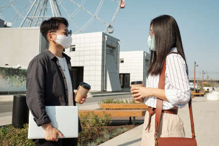 Two young colleagues in protective masks looking at each other while having drinks and communicating...