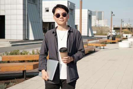 Young Chinese woman in casualwear and sunglasses holding laptop and drink while spending time in modern parkの写真素材