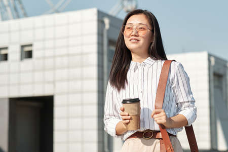 Young smiling businesswoman with drink and handbag standing in front of camera on sunny day against modern architectureの写真素材